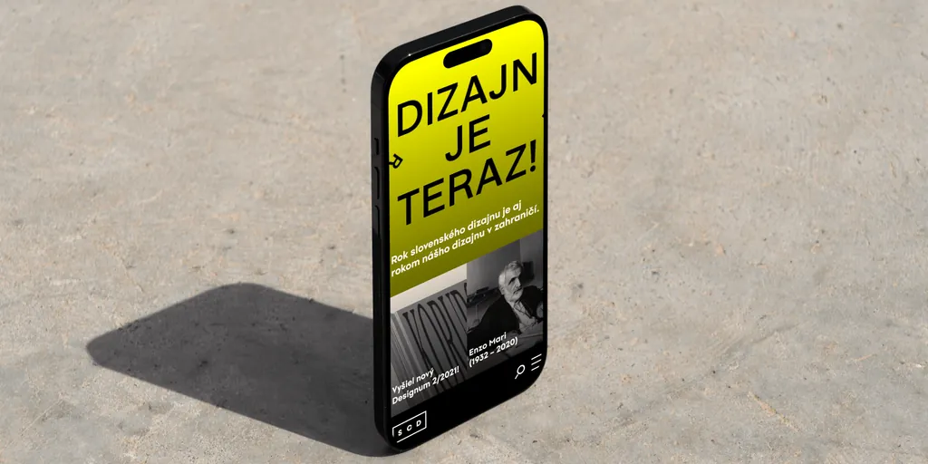 The SCD website on a phone standing on concrete: “Dizajn je teraz!” (design is now)
