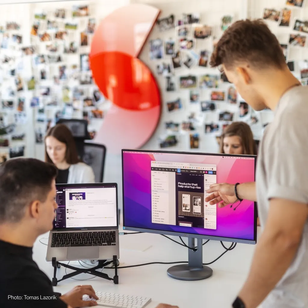 Sudolabs developers at a monitor, the red S mark on the wall behind them