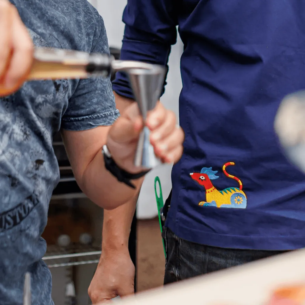 The La Luz alebrije embroidered on a navy La Hacienda staff shirt, as a drink is poured into a jigger