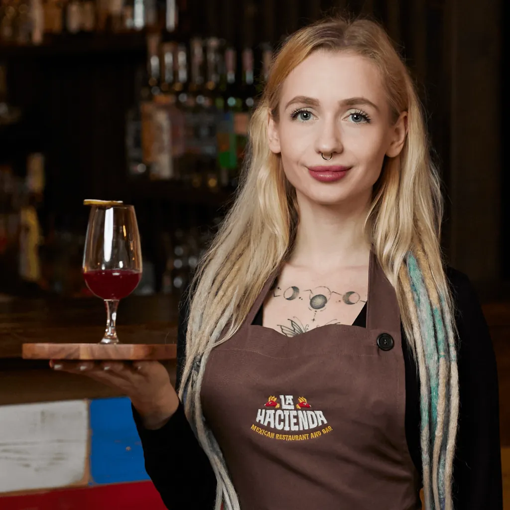 A La Hacienda server in a brown apron with the embroidered logo, holding a drink on a wooden tray